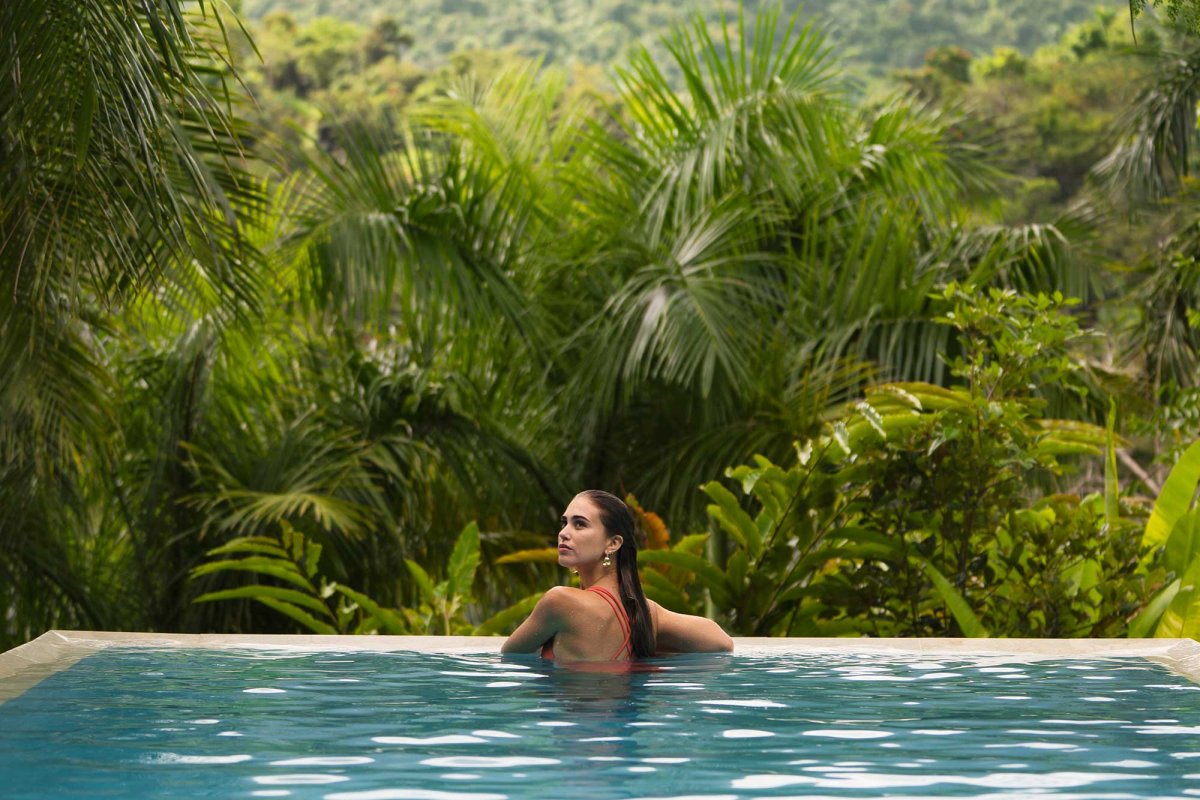 A woman rests on the side of an infinity pool overlooking a lush forest in the mountains of Puerto Rico.