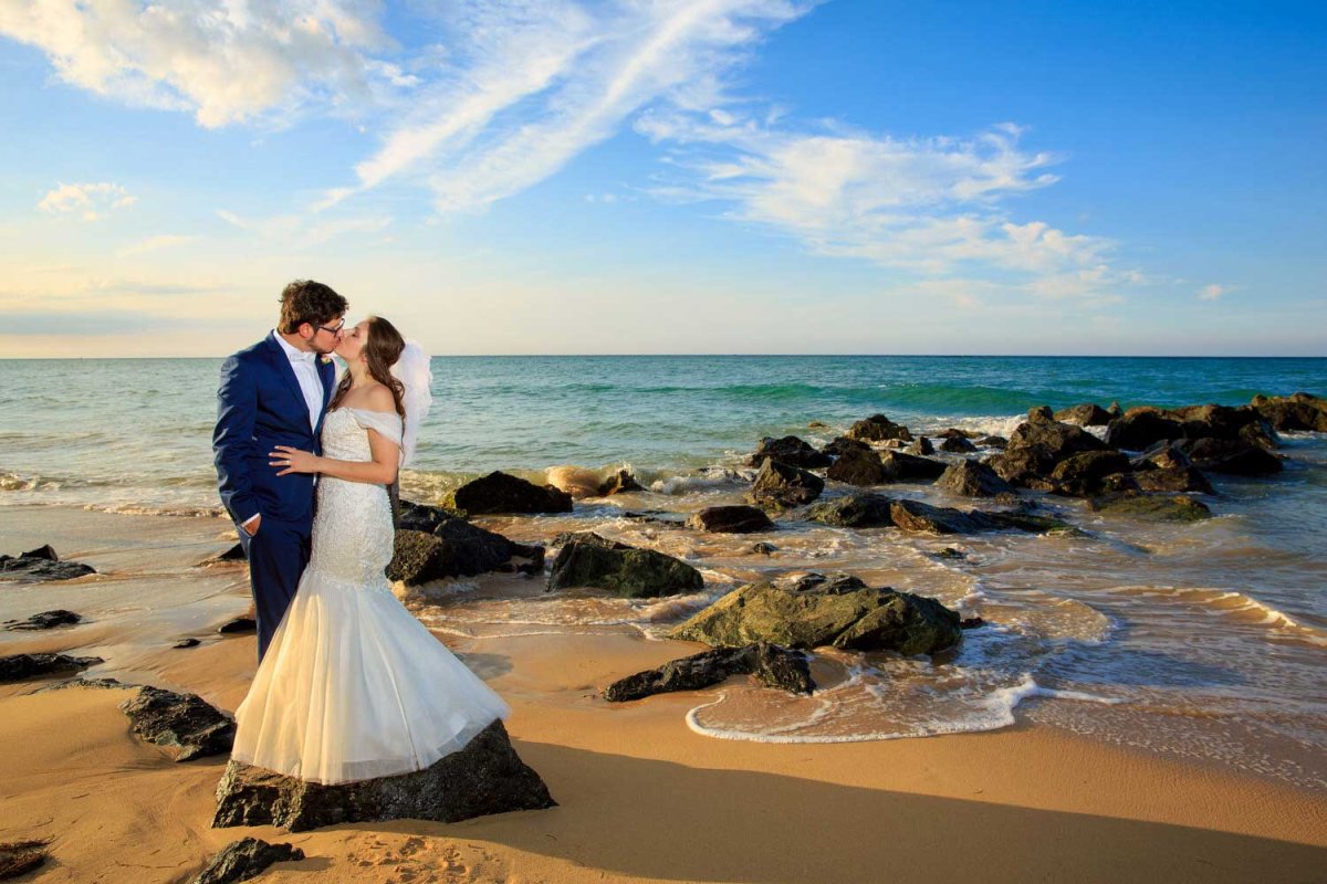 Una novia y un novio recién casados se besan mientras están de pie en una playa salpicada de rocas en Puerto Rico. Foto de Noel Del Pilar.