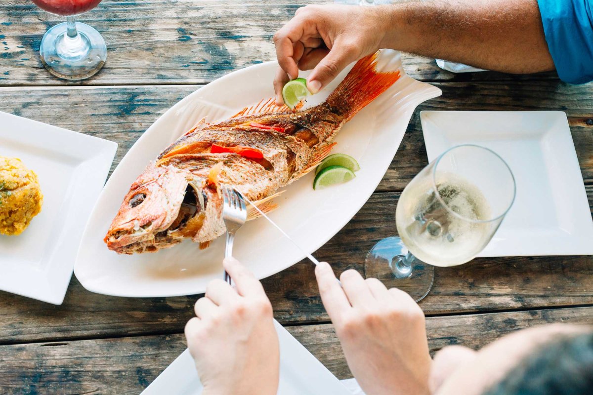 Overhead view of a plate of whole fish and a glass of wine, with one pair of hands holding a knife and fork and another hand squeezing a lime slice. Salpicón restaurant, Arecibo, Puerto Rico.