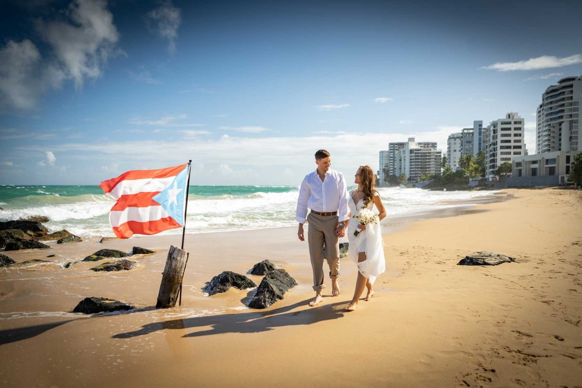 A bride and groom walk on the beach near the San Juan Marriott Resort & Stellaris Casino, with a Puerto Rican flag planted in the sand.