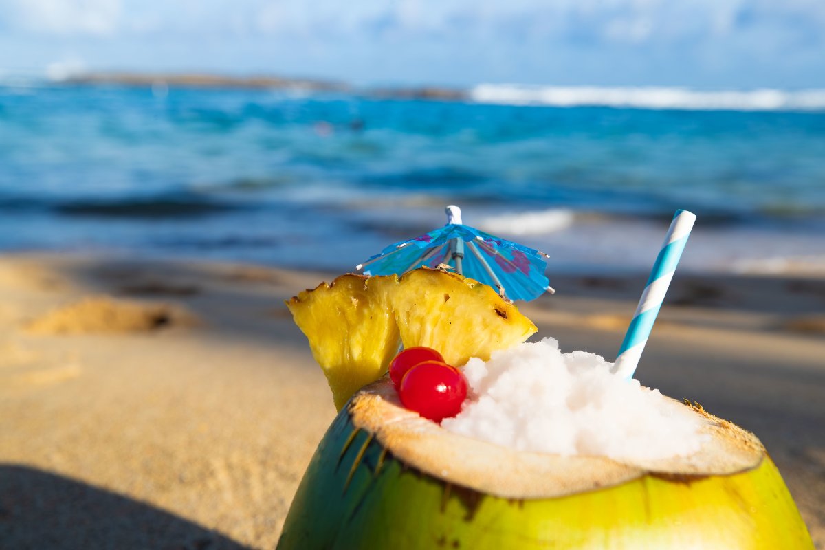 Photo of coconut filled with piña colada with a beach in the background.