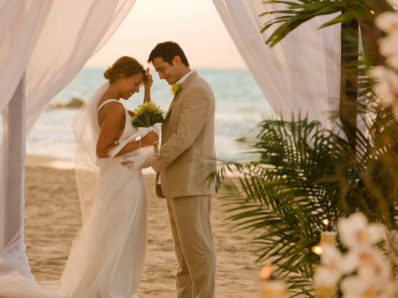 Una novia y un novio se enfrentan frente a un altar cubierto con tela blanca en una playa de Puerto Rico.