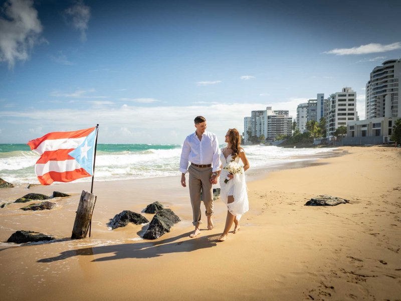 Una novia y un novio caminan por la playa cerca del San Juan Marriott Resort & Stellaris Casino, con una bandera puertorriqueña plantada en la arena.