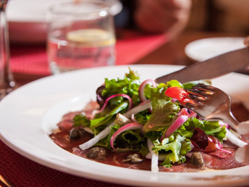 A person cutting a plate of salad greens on top of thinly sliced beef at Restaurante Alexandra.