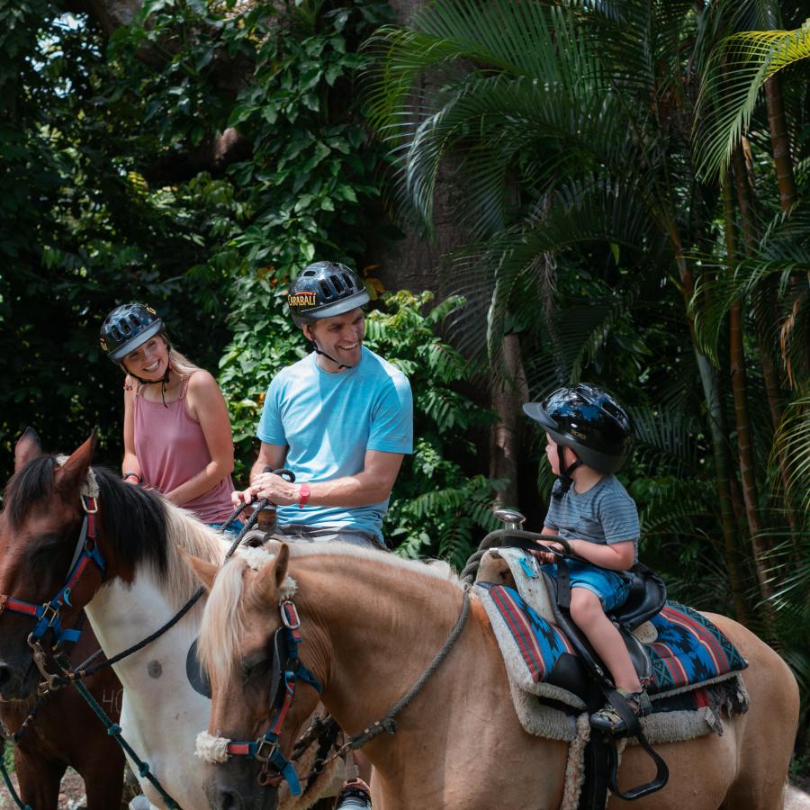 A famiy of three sitting atop three horses in a green outdoor space.