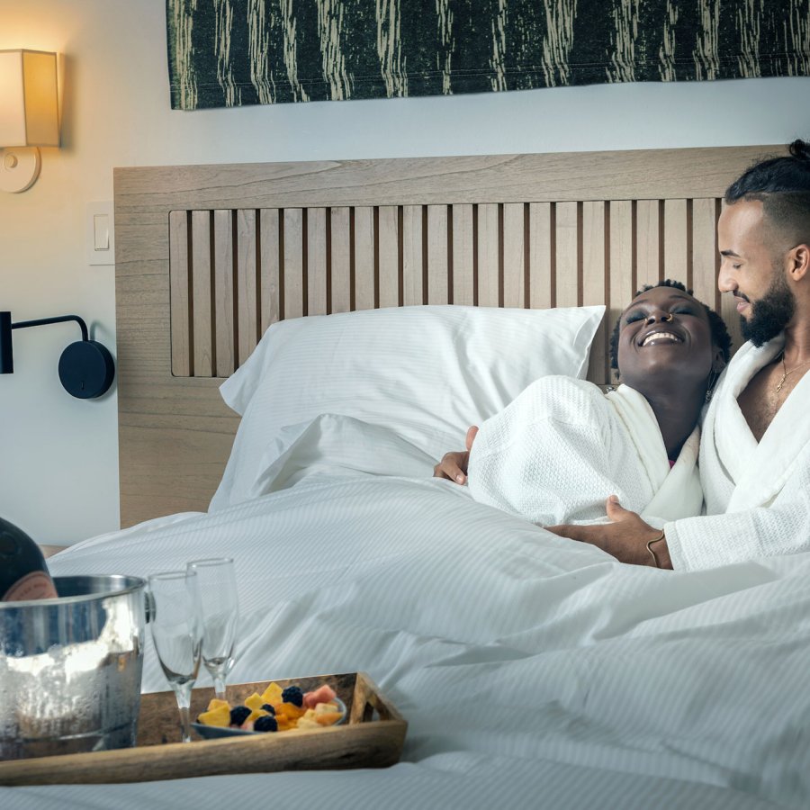 A couple lies in a hotel bed wearing white robes with a bottle of wine at the Hotel Palacio Provincial in San Juan, Puerto Rico.