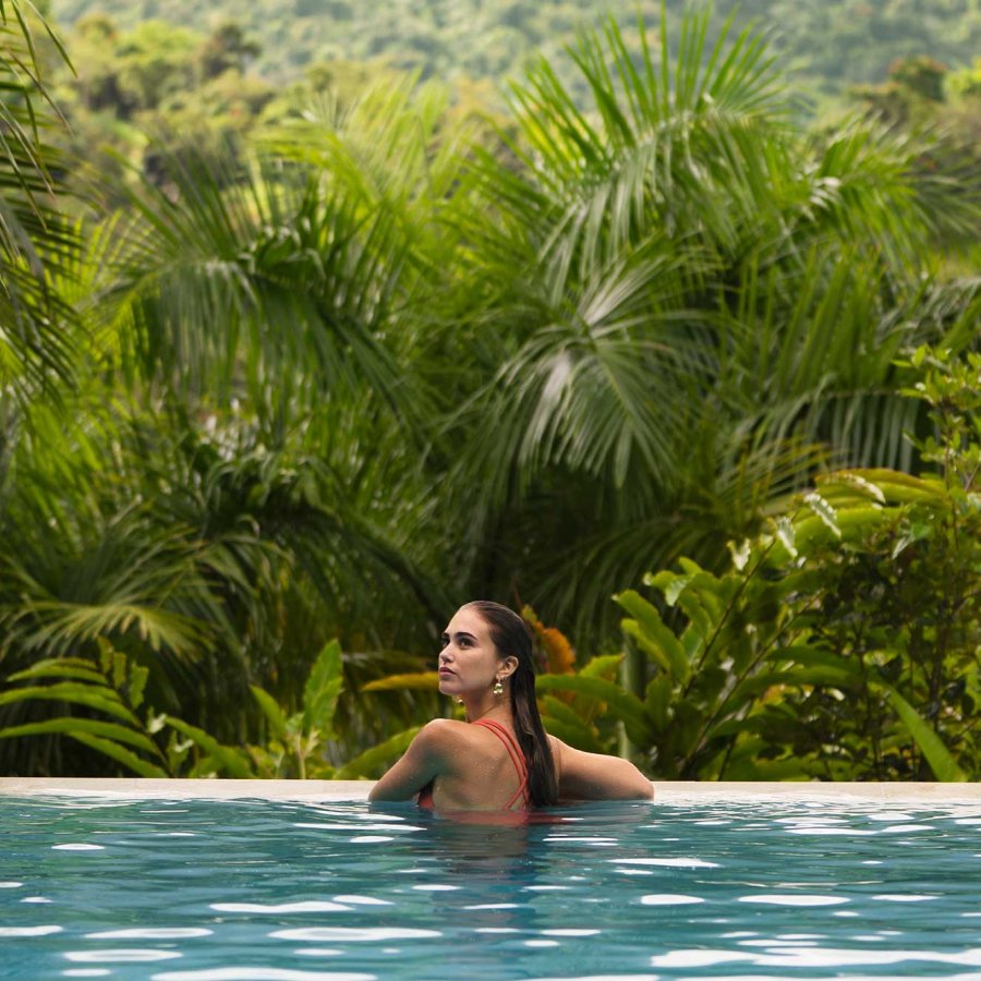 A woman rests on the side of an infinity pool overlooking a lush forest in the mountains of Puerto Rico.